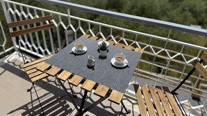 Un balcon avec une table dressée pour deux personnes, comprenant une table avec un plateau en métal grillagé, deux chaises en bois et une balustrade blanche.