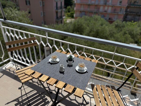 A balcony with a table set for two, featuring a metal grill table, two wooden chairs, and a white railing.