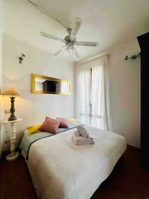 Une chambre à coucher avec un lit, un ventilateur de plafond, une lampe de chevet, un miroir et une fenêtre avec des rideaux.