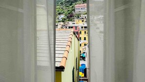 A view through a window with sheer curtains, revealing a balcony with a tiled roof and a building with multiple stories in the background.