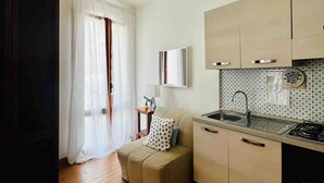 A compact kitchen with a sink, stove, and cabinets, a small seating area with a cushion, and a window with sheer curtains.