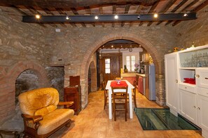 A stone-walled room with a fireplace, a dining table with chairs, and a kitchen area visible through an archway.