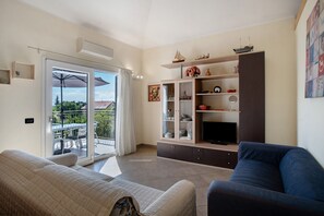 Un soggiorno con divano, televisione, mobile contenitore e un balcone con tavolo e ombrellone.