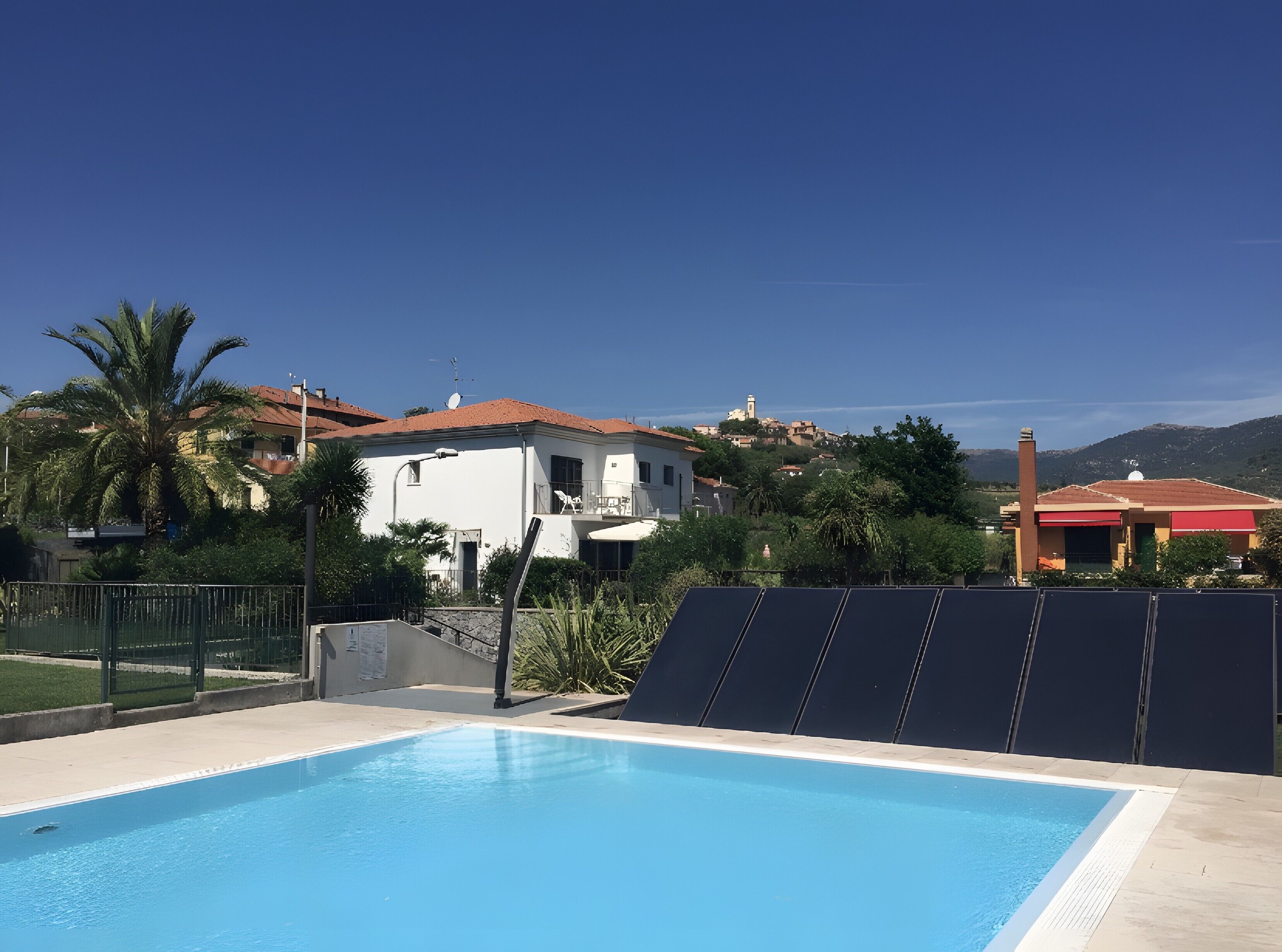 A swimming pool with a clear blue sky, a white building with a red roof, and a red-roofed structure in the background.