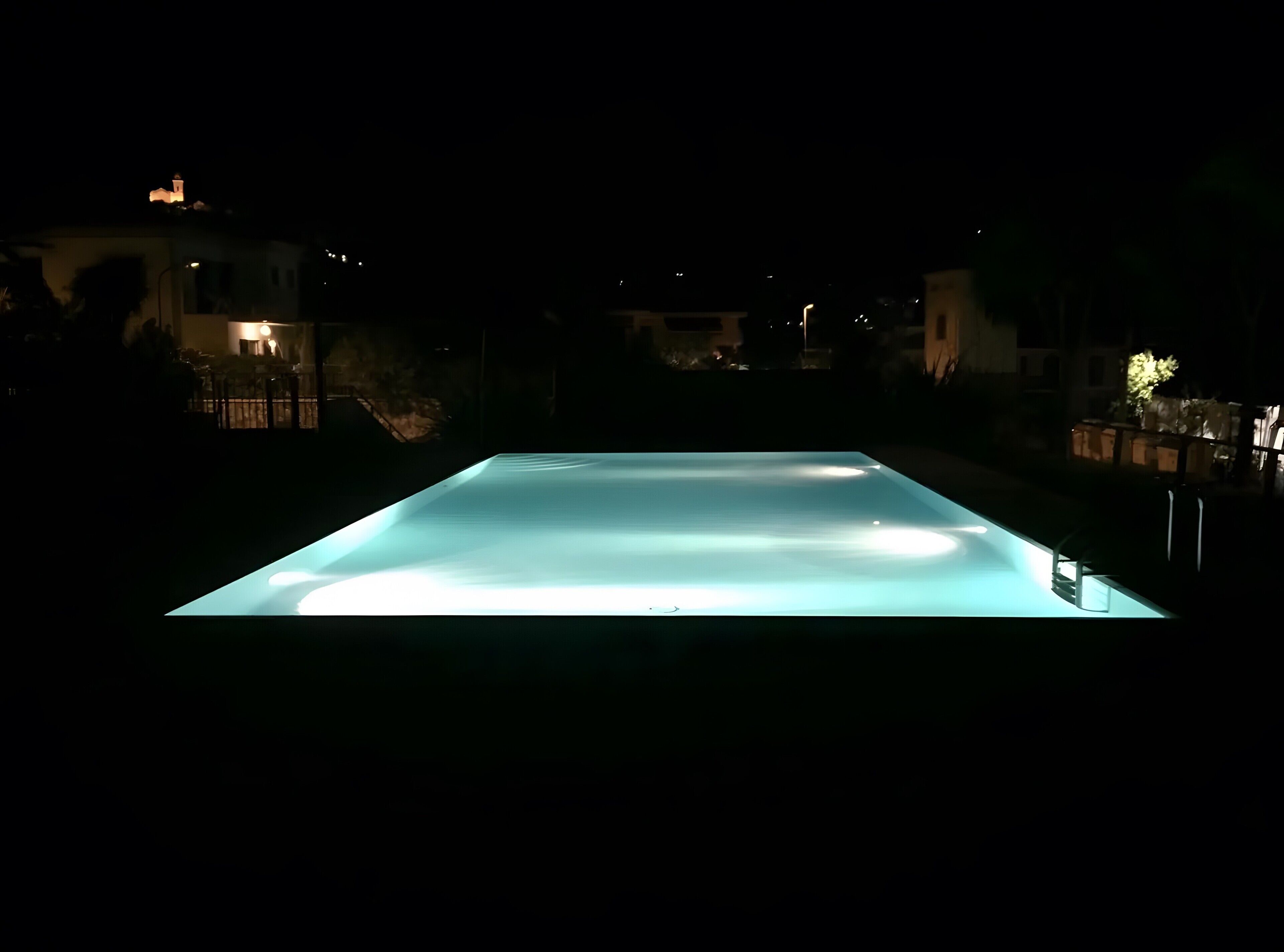 Una piscina rectangular iluminada de noche, rodeada de edificios y una valla.