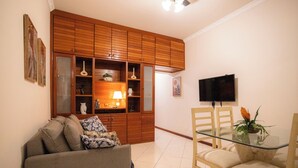 A living room with a sofa, a glass-top dining table, wooden cabinets, and a television. - SUH Copacabana 71 Princess Beach (Rio de Janeiro)