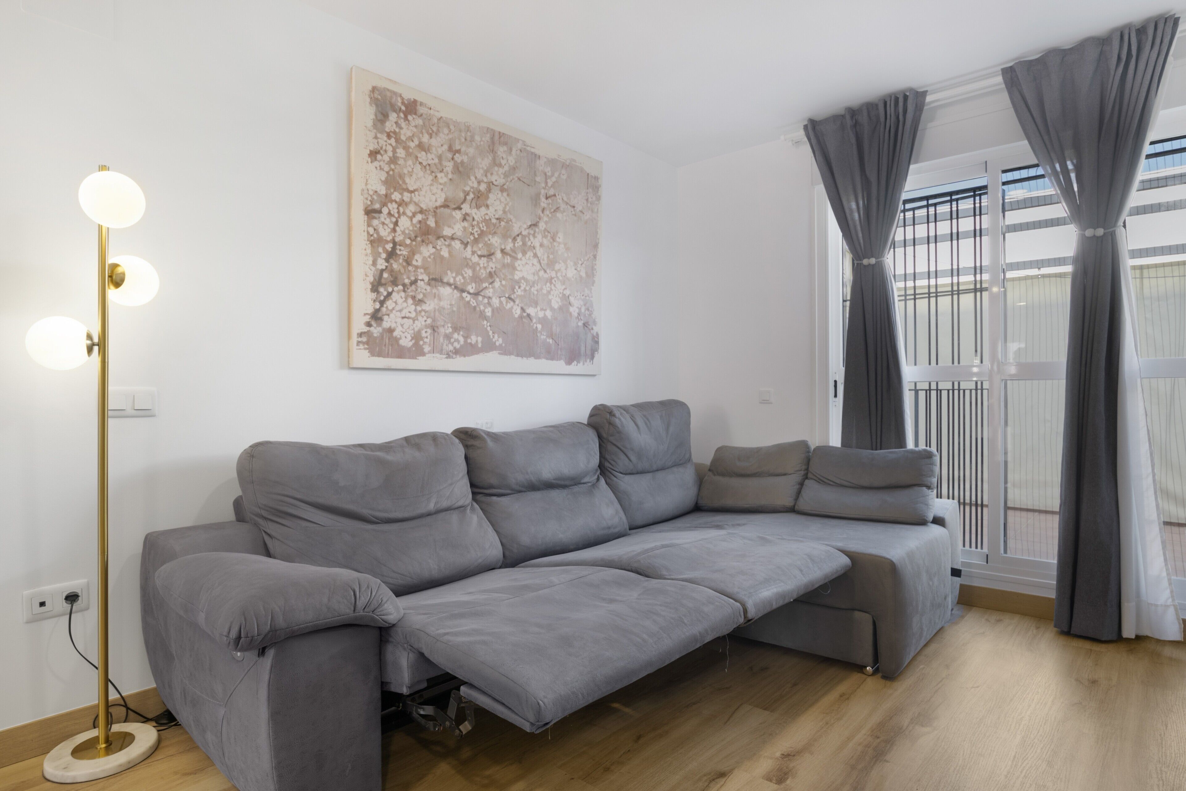 A modern living room with a grey sectional sofa, a floor lamp, a large abstract painting, and a balcony with grey curtains.