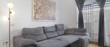 A modern living room with a grey sectional sofa, a floor lamp, a large abstract painting, and a balcony with grey curtains.