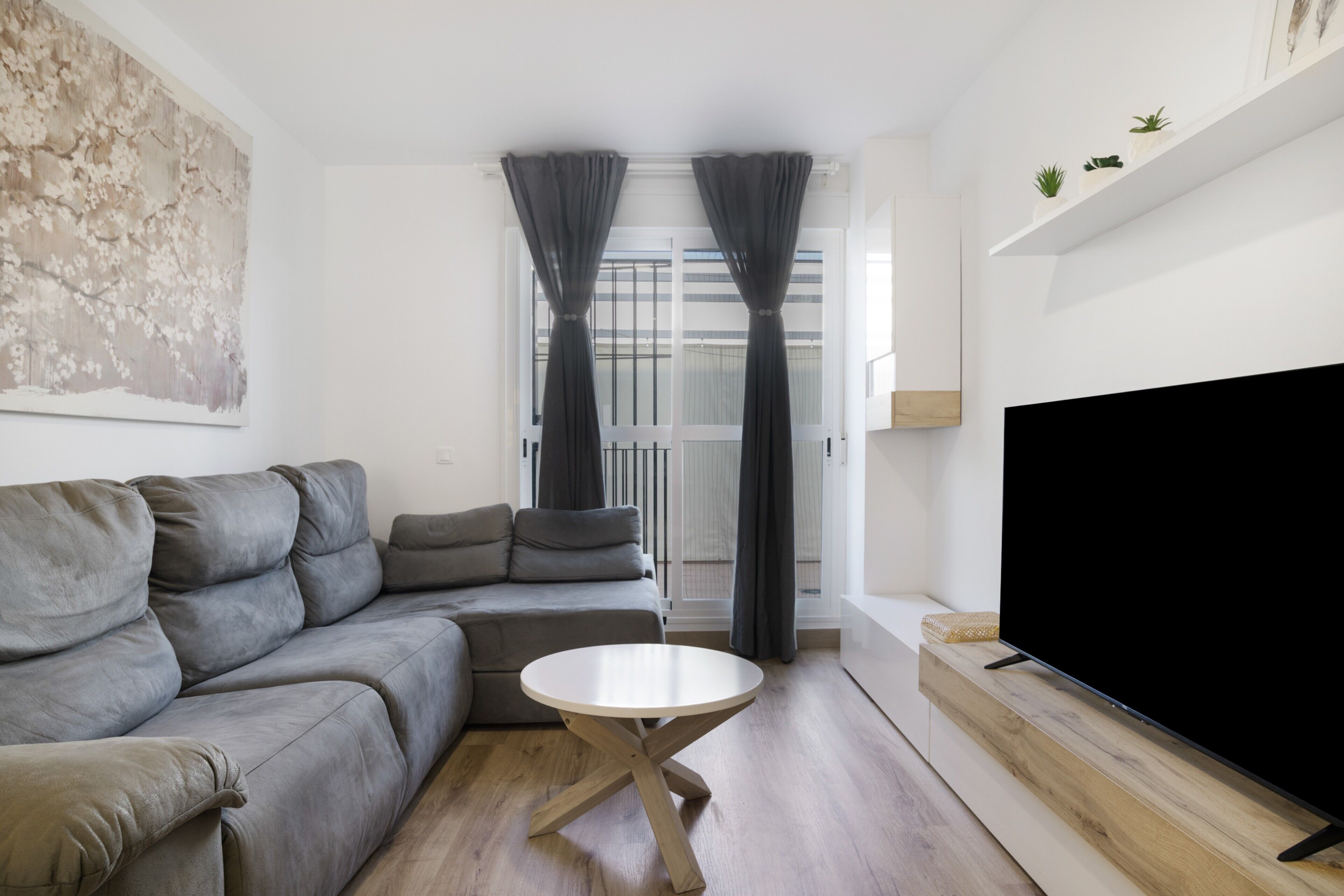 Ein modernes Wohnzimmer mit einem grauen Ecksofa, einem runden Couchtisch aus Holz, einem Flachbildfernseher auf einem Holzständer und einem großen abstrakten Gemälde an der Wand.