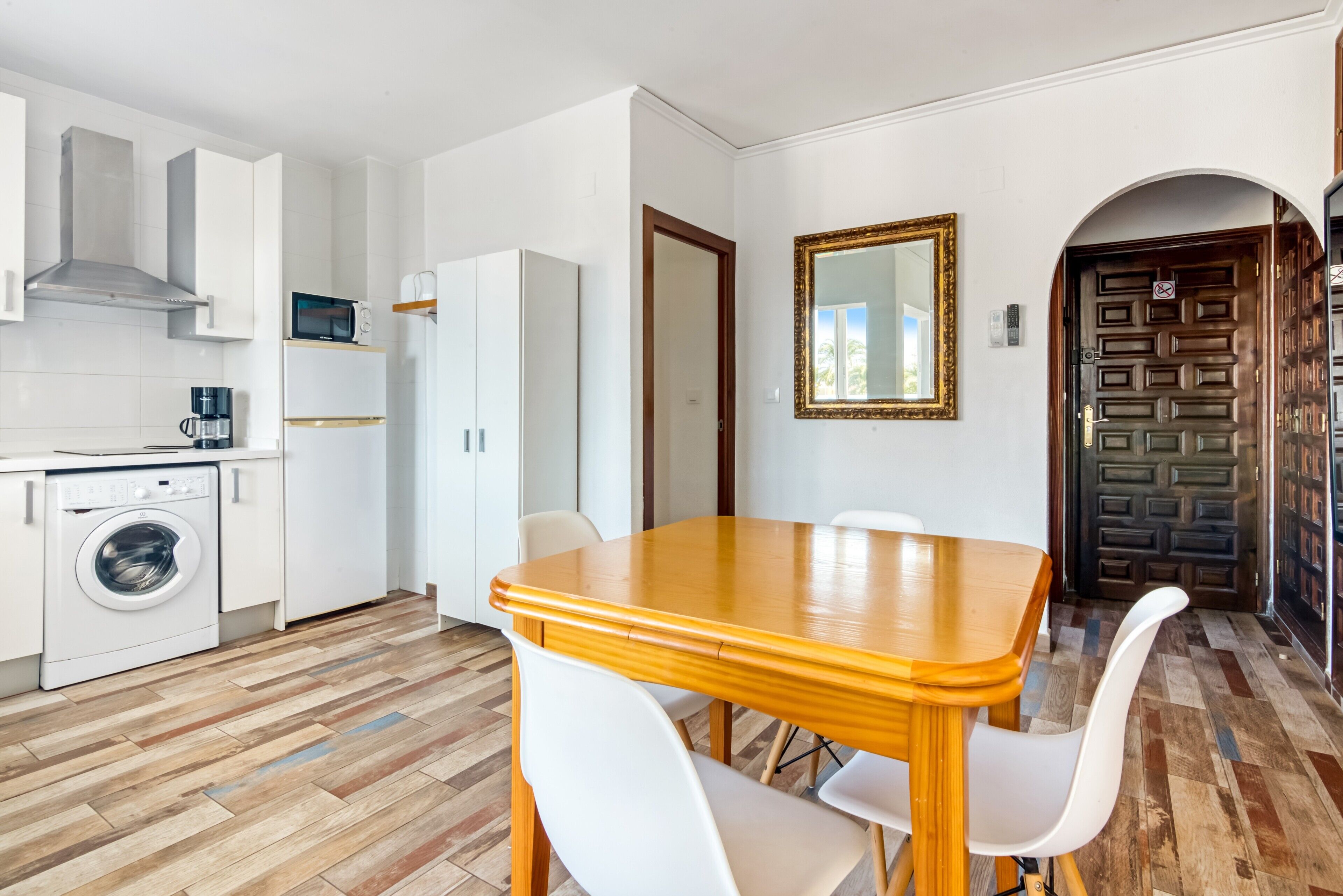 A compact kitchen with a washing machine, a microwave, and a coffee maker. There is a wooden dining table with white chairs.