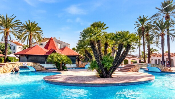 A swimming pool with palm trees, a stone bridge, and a building with a red roof. - Apartment 'El Palmar' with Sea View, Wi-Fi and Air Conditioning (Denia)