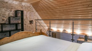 A bedroom with a wooden ceiling, a stone wall, a bed, a metal ladder, and a small shelf with a statue. - Le Portougais, Chanaz – Apartment with Wi-Fi and Air Conditioning (Chanaz)