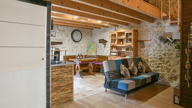 A cozy living room with a wooden ceiling, a stone wall, a sofa with patterned cushions, a wooden cabinet, and a dining table with chairs. - Le Portougais, Chanaz – Apartment with Wi-Fi and Air Conditioning (Chanaz)
