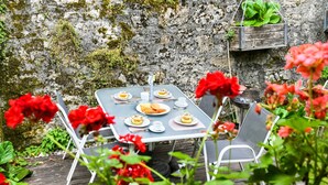 A table set outdoors with plates of food, surrounded by white chairs, against a stone wall backdrop. - Le Portougais, Chanaz – Apartment with Wi-Fi and Air Conditioning (Chanaz)