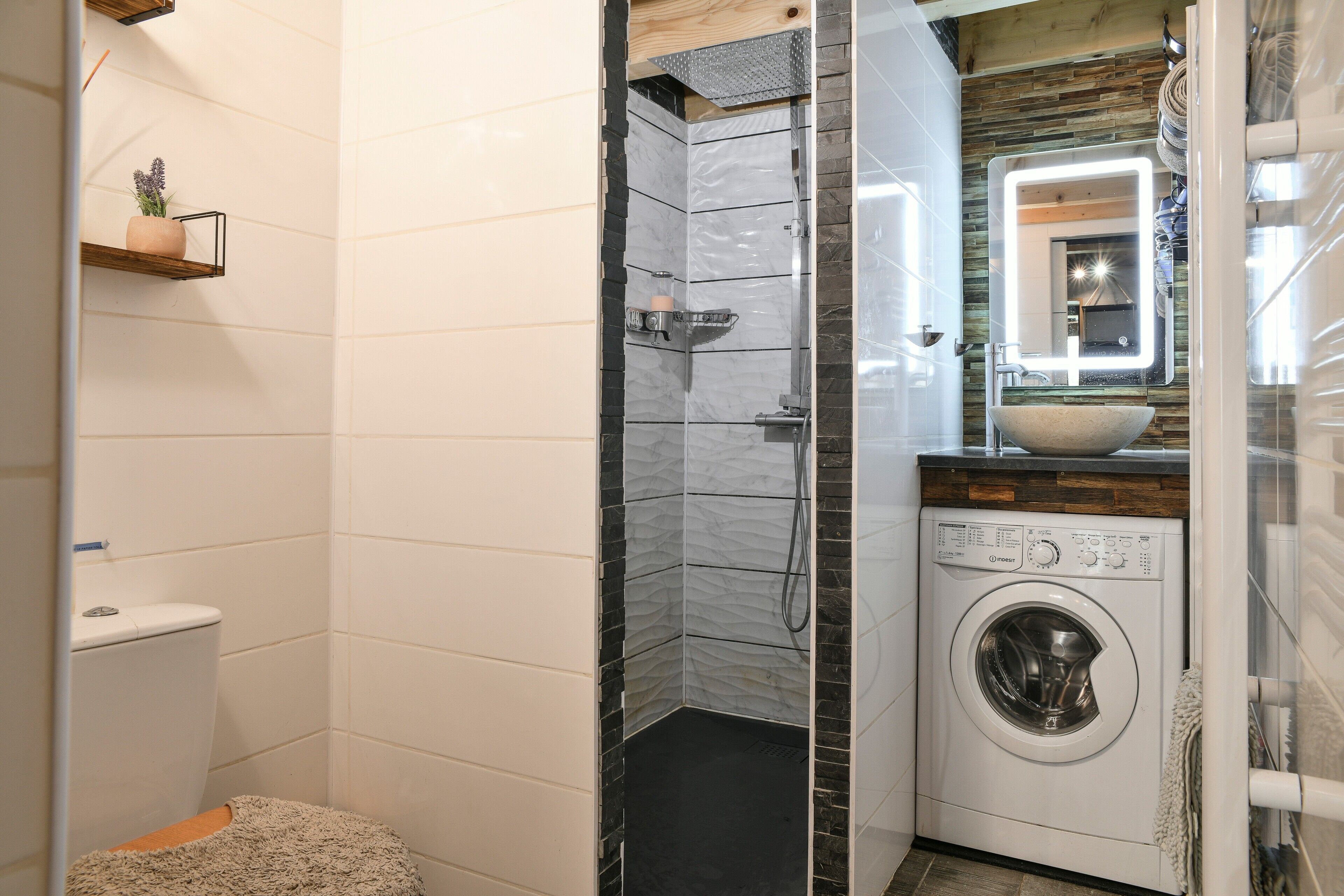Un baño compacto con ducha, inodoro, lavadora y una pequeña estantería con una planta.