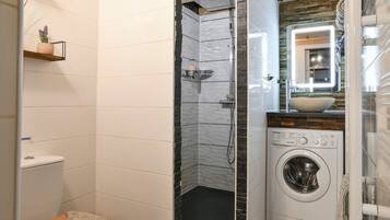 Un baño compacto con ducha, inodoro, lavadora y una pequeña estantería con una planta.