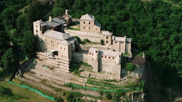 Eine Vogelperspektive auf einen historischen Gebäudekomplex aus Stein mit mehreren Bauten, umgeben von üppiger Vegetation und einer steilen Klippe.