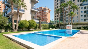 A swimming pool with a tiled deck, surrounded by palm trees and a multi-story building in the background. - 'Boliche' Apartment with Shared Pool, Wi-Fi, and Air Conditioning (Alicante (Alacant))
