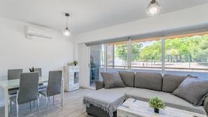 A modern living room with a sofa, a dining table with chairs, and a large window with a view of greenery. - 'Boliche' Apartment with Shared Pool, Wi-Fi, and Air Conditioning (Alicante (Alacant))