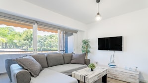 A modern living room with a sectional sofa, a coffee table, a TV mounted on the wall, and a view of greenery outside. - 'Boliche' Apartment with Shared Pool, Wi-Fi, and Air Conditioning (Alicante (Alacant))