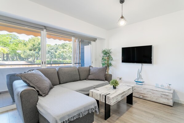 A modern living room with a sectional sofa, a coffee table, a TV mounted on the wall, and a view of greenery outside.