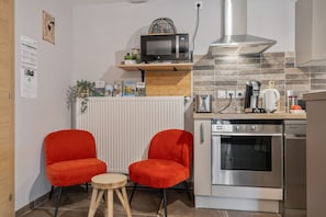 Une cuisine compacte équipée d’un four à micro-ondes, d’un four et d’une machine à café. On y trouve deux chaises rouges et une petite table en bois.