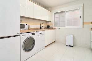 Una cocina moderna con armarios blancos, microondas, cafetera, lavadora y contenedor de basura.