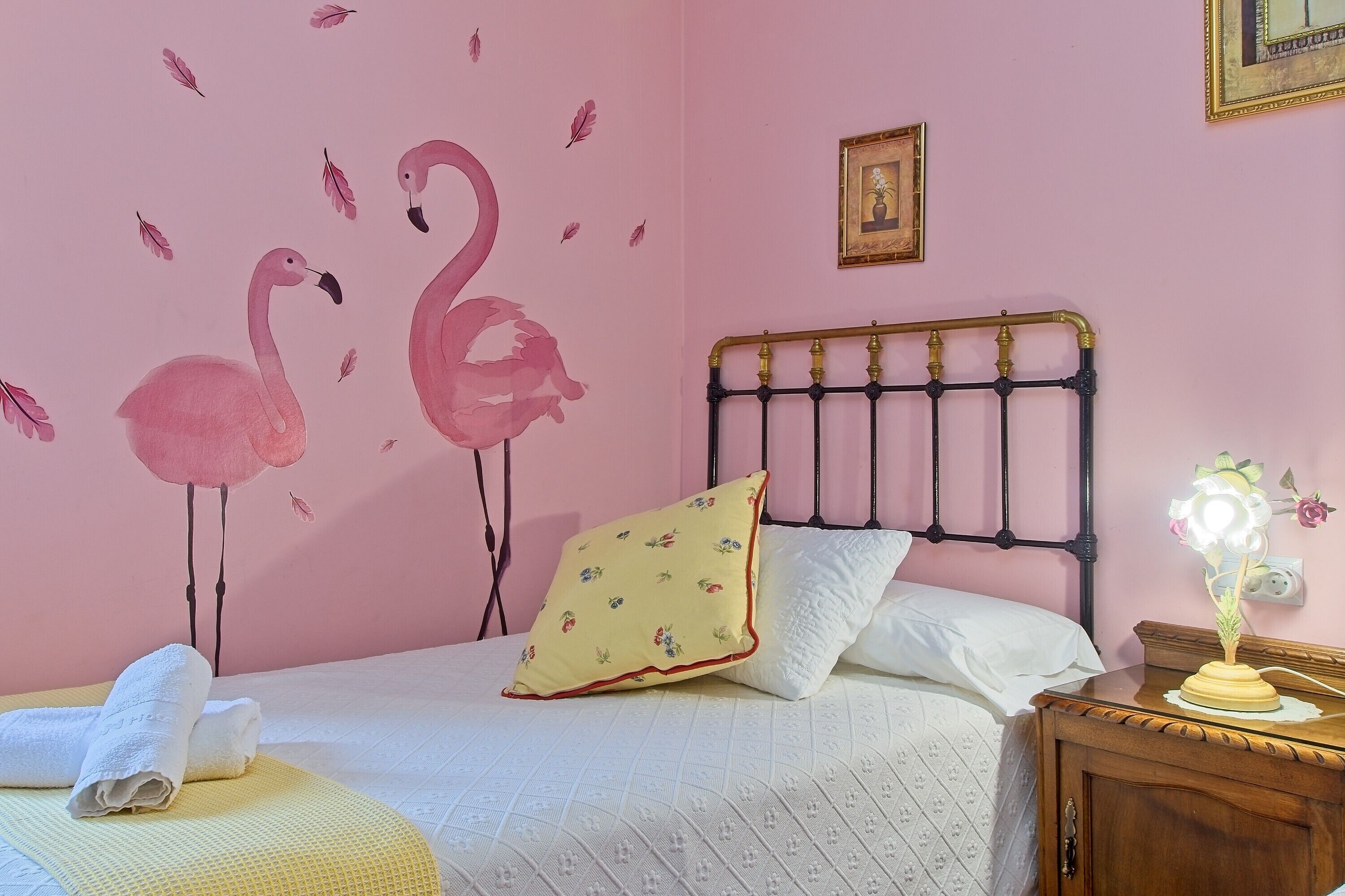 Een slaapkamer met een roze wand, een bed met witte beddengoed, een bloemenkussen, een nachtkastje met een lamp, en een flamingo-wandsticker.