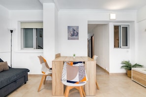 A modern living room with a sofa, a dining table, and chairs. There is a window with blinds, a painting on the wall, and a plant on the floor.