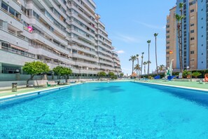 Une grande piscine avec des chaises longues, un bar et un restaurant, situés à côté d’un immeuble d’hôtel à plusieurs étages.