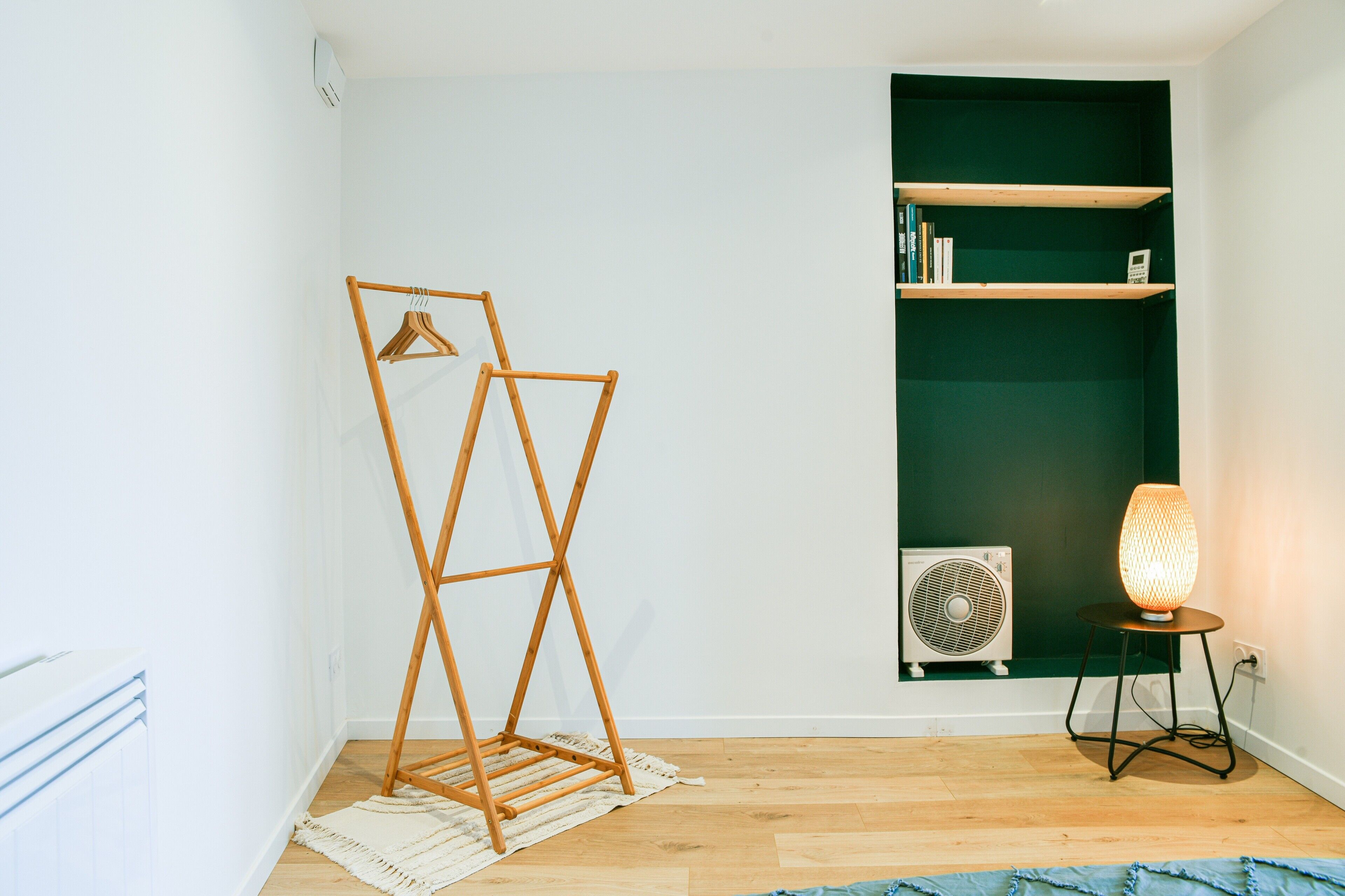 Ein Zimmer mit einem Wäscheständer aus Holz, einem Standventilator und einem Tisch mit einer Lampe.