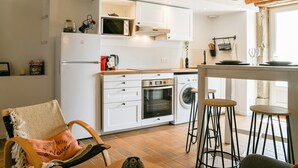 Una cocina con armarios blancos, microondas, horno y lavadora. Hay taburetes y una silla con cojín.