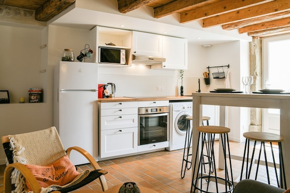 A kitchen with white cabinets, a microwave, oven, and a washing machine. There are stools and a chair with a cushion.
