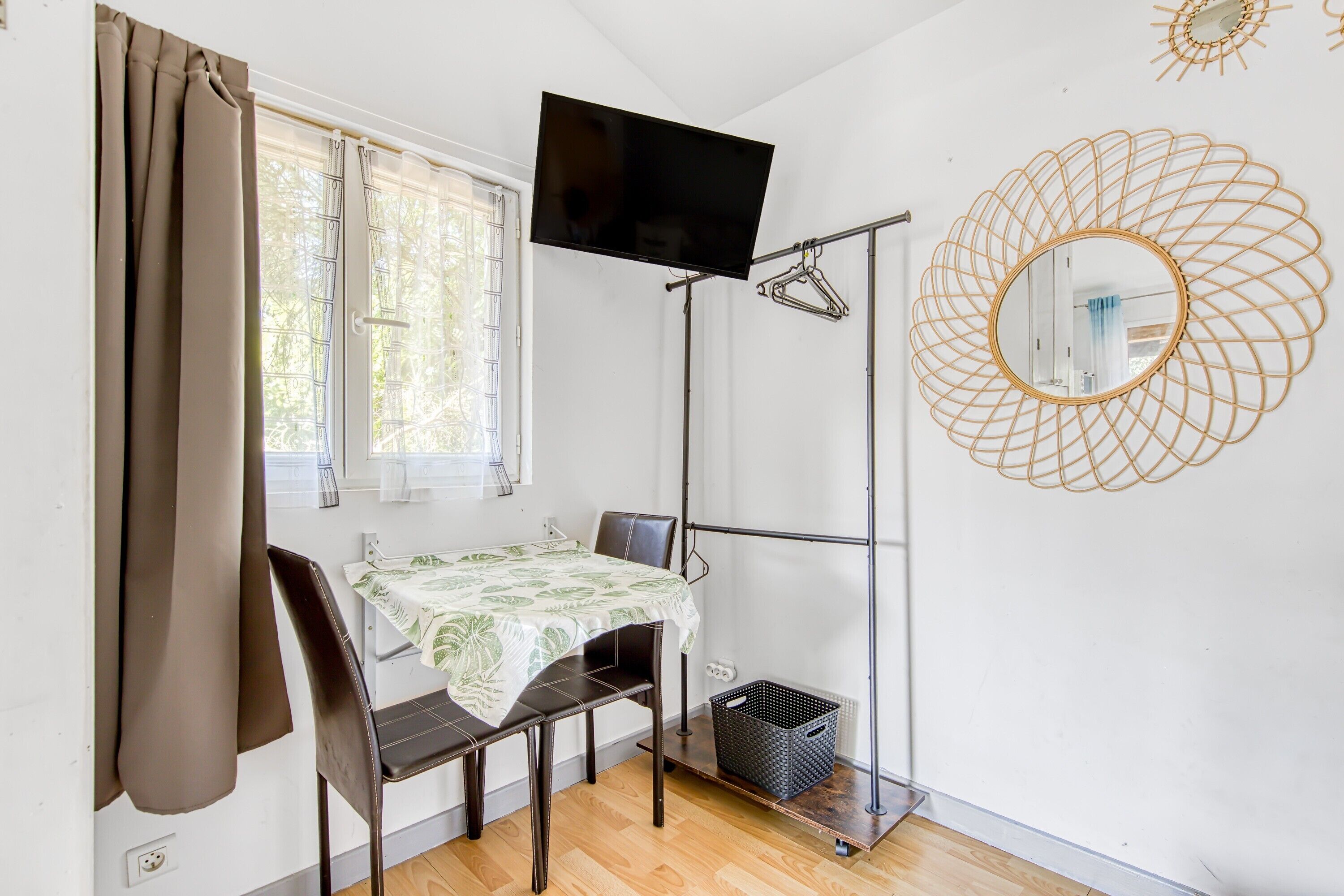 Une pièce comprenant un lit, une table recouverte d’une nappe, une chaise, un téléviseur fixé au mur, une fenêtre avec des rideaux et un miroir décoratif.