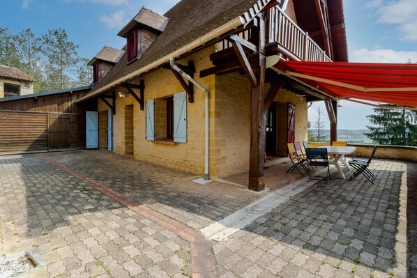 A rustic building with a balcony, a red awning, and a paved patio area.