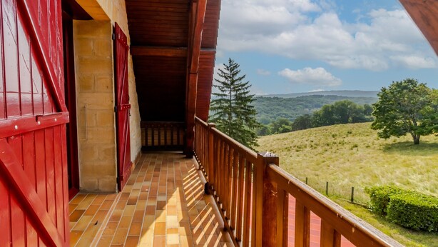 Un balcón de madera con contraventanas rojas, suelo de baldosas y vistas a un paisaje montañoso.