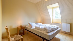 Un dormitorio con cama alta, una silla de madera, una mesita de noche con lámpara y una ventana con vista a árboles.