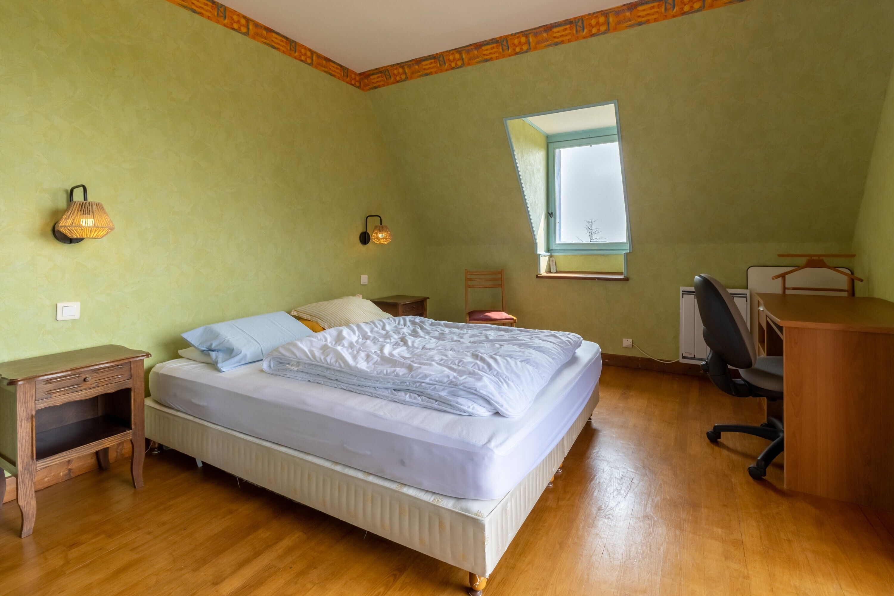 A bedroom with a bed, bedside tables, a desk, a chair, a window, and a wall-mounted lamp.
