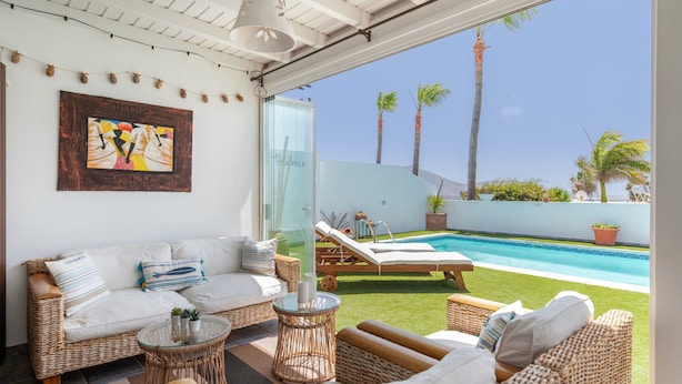 A sunlit room with a sofa, lounge chairs, a glass table, and a view of a swimming pool and palm trees.