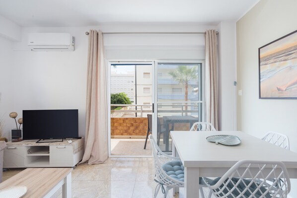 Un salón moderno con televisor de pantalla plana, una mesa de comedor y un balcón con vistas a edificios y palmeras.
