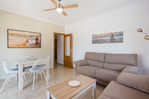 A living room with a beige sofa, a dining table with white chairs, a ceiling fan, and two wall-mounted paintings.