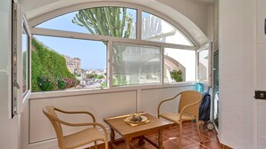 A room with a large window, two wicker chairs, a small table, and a view of buildings and greenery outside.