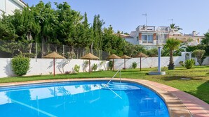 A swimming pool with a clear blue water, surrounded by a white wall, and a grassy area with trees and a fence in the background.