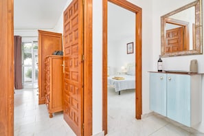 Un pasillo con puertas de madera, una cama con colcha blanca, una mesita de noche y un armario con espejo.