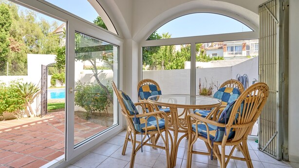 A sunlit patio with a round table and wicker chairs, a glass door leading outside, and a view of a swimming pool and greenery.