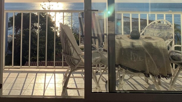 A balcony with a view of the ocean and a sunset. There are two wicker chairs and a table with a cloth.