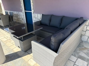A wicker sofa with cushions, a glass-top table, and a blue door in a stone-walled room.