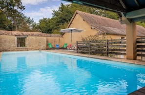 A swimming pool with a wooden deck, a covered area, and a building with a tiled roof in the background. - Le Pigeonnier – Characterful House with Garden and Shared Pool in Dordogne (Mayac)