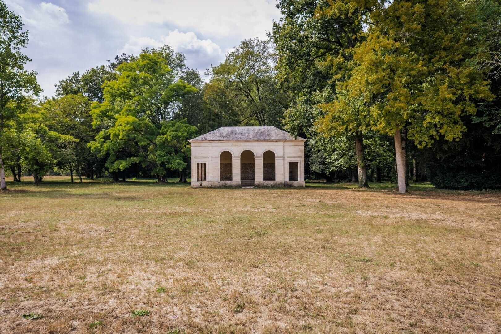 Un bâtiment historique doté d’une entrée centrale en arc de cercle, entouré d’un champ verdoyant et d’arbres.
