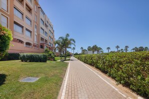 A pathway leading to a multi-story building with balconies, surrounded by well-maintained hedges and palm trees.